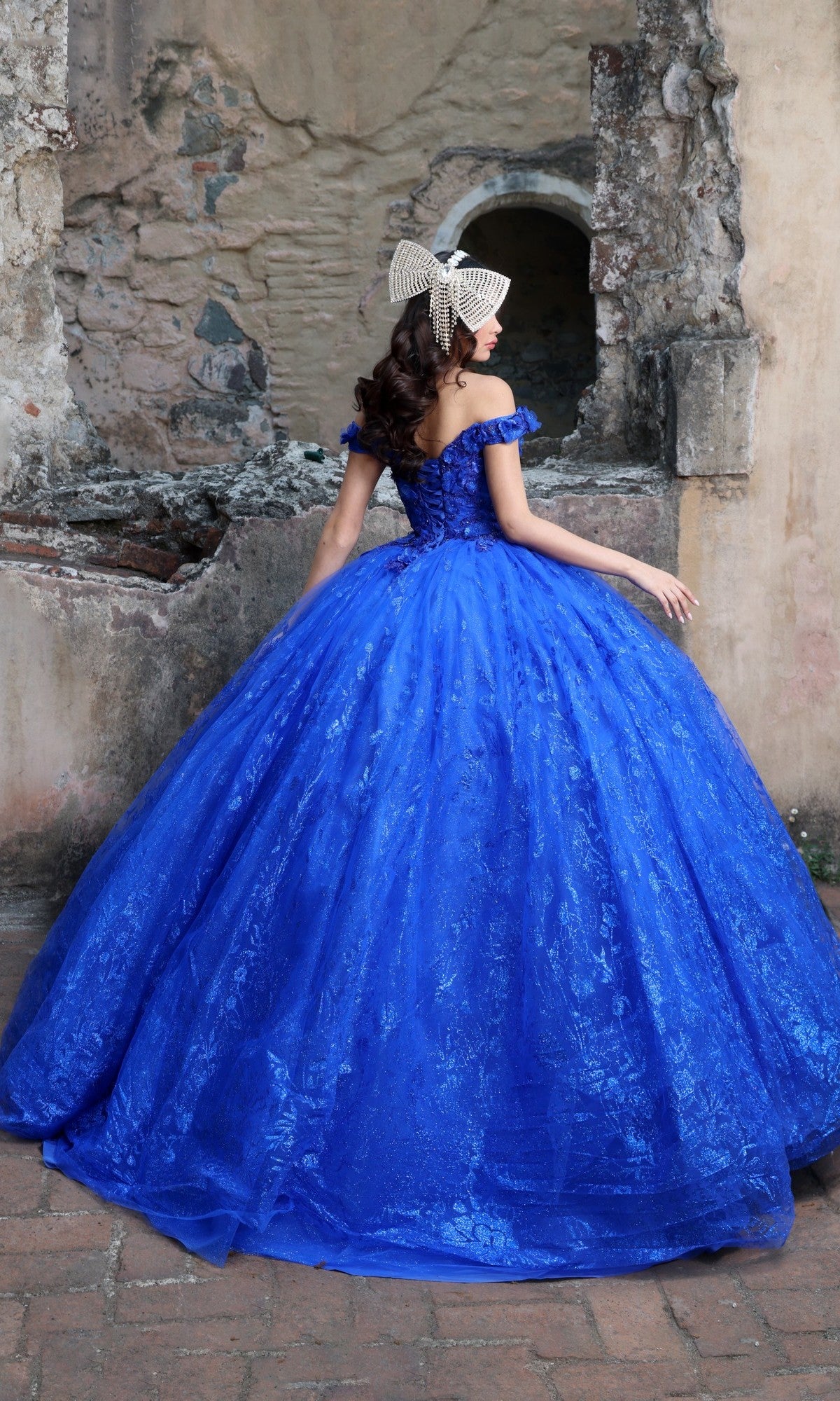 Someone wearing the Quinceanera Dress Q129 By Nina Canacci and a decorative headpiece stands with their back to the camera in front of an old stone wall.